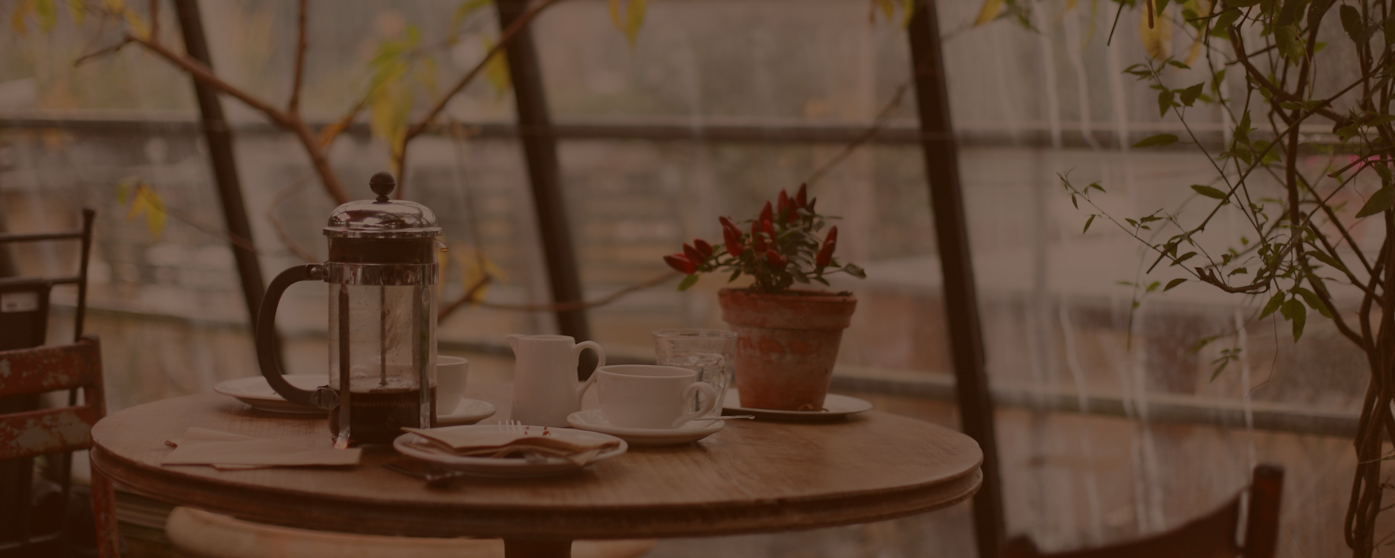 Café setting with a French press, cups, and saucers on a wooden table with a blurred background.